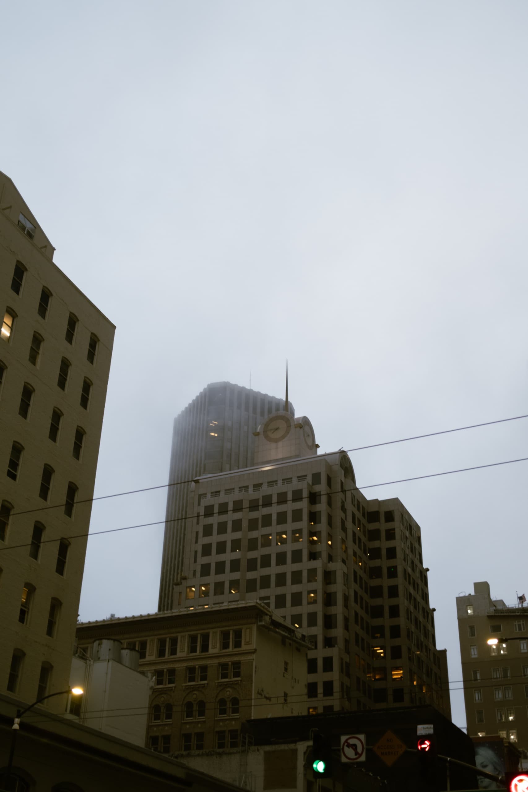 Foggy downtown street canyon at dusk with a clock-faced building and a green traffic light.