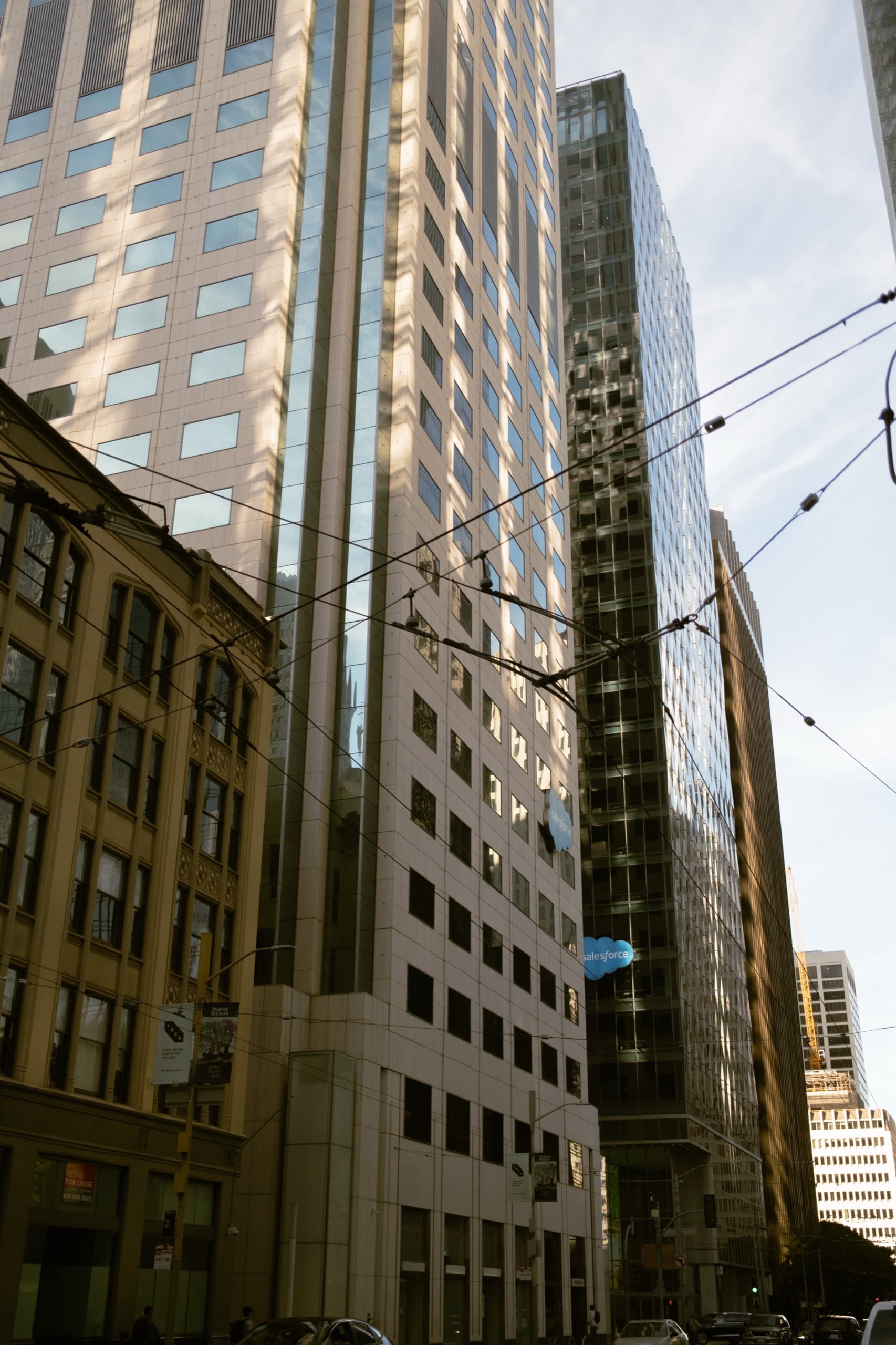 Low-angle daytime street canyon with a white tower, trolley wires, and a FOR LEASE sign.