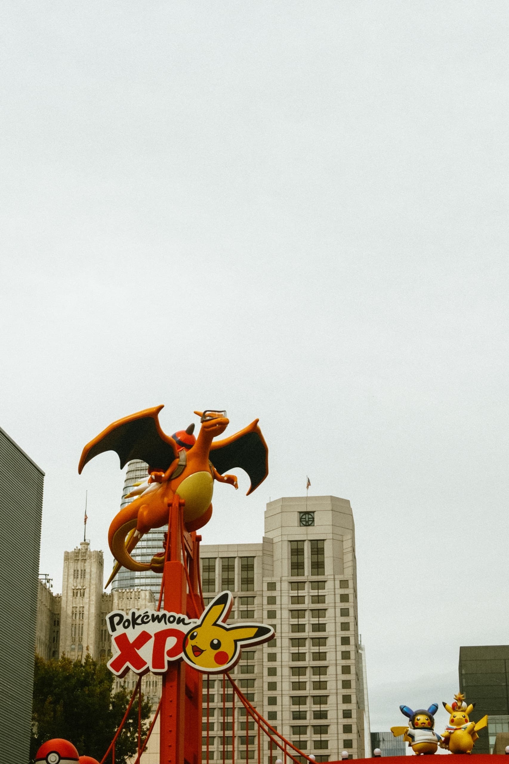 Charizard statue with goggles on a red tower, two costumed Pikachu, city skyline.
