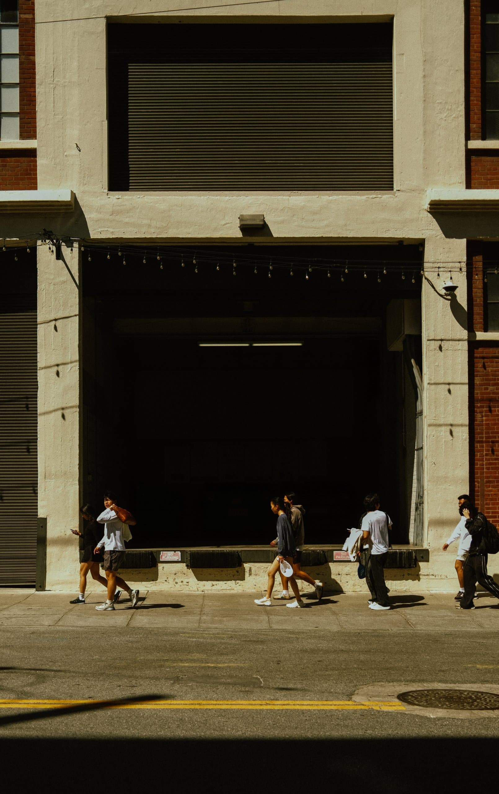 People walking on a sunlit sidewalk in front of a beige industrial loading-dock opening with a string of bulbs, long shadows on the pavement.